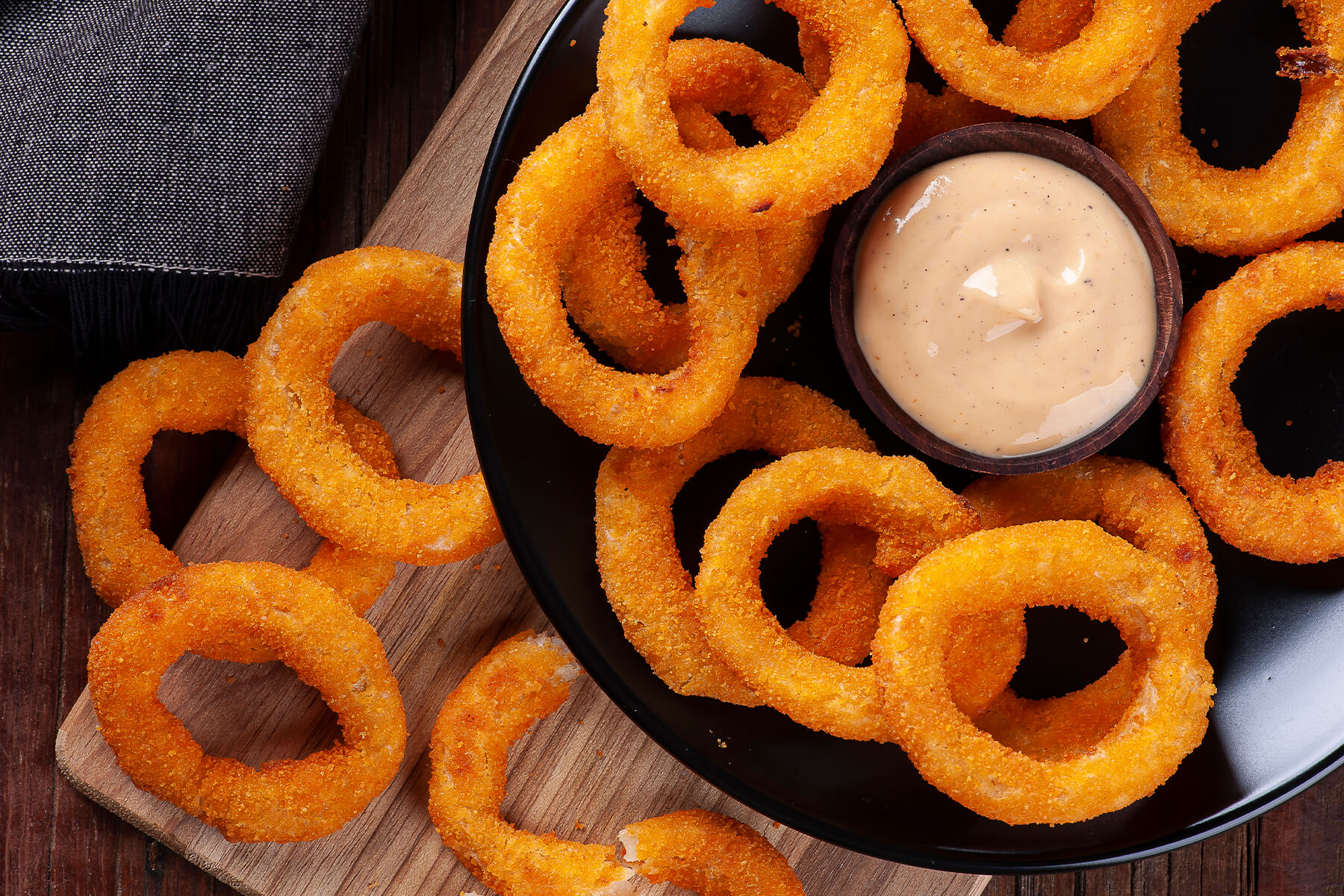 Nahaufnahme goldbraun frittierter Zwiebelringe auf einem schwarzen Teller mit einem Schälchen cremiger scharfer Dip-Sauce