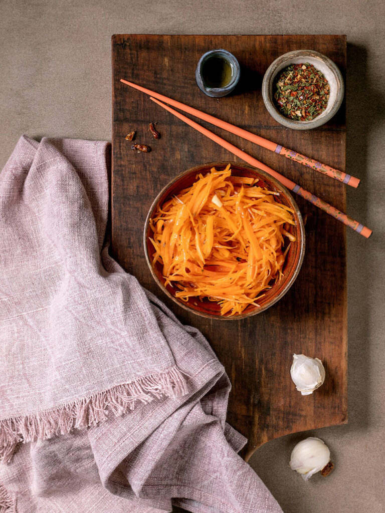 Koreanischer Karottensalat aus langen, glänzend marinierten Karottenstreifen in einer Keramikschale auf einem dunklen Holzbrett, daneben Stäbchen, Gewürze und Knoblauch