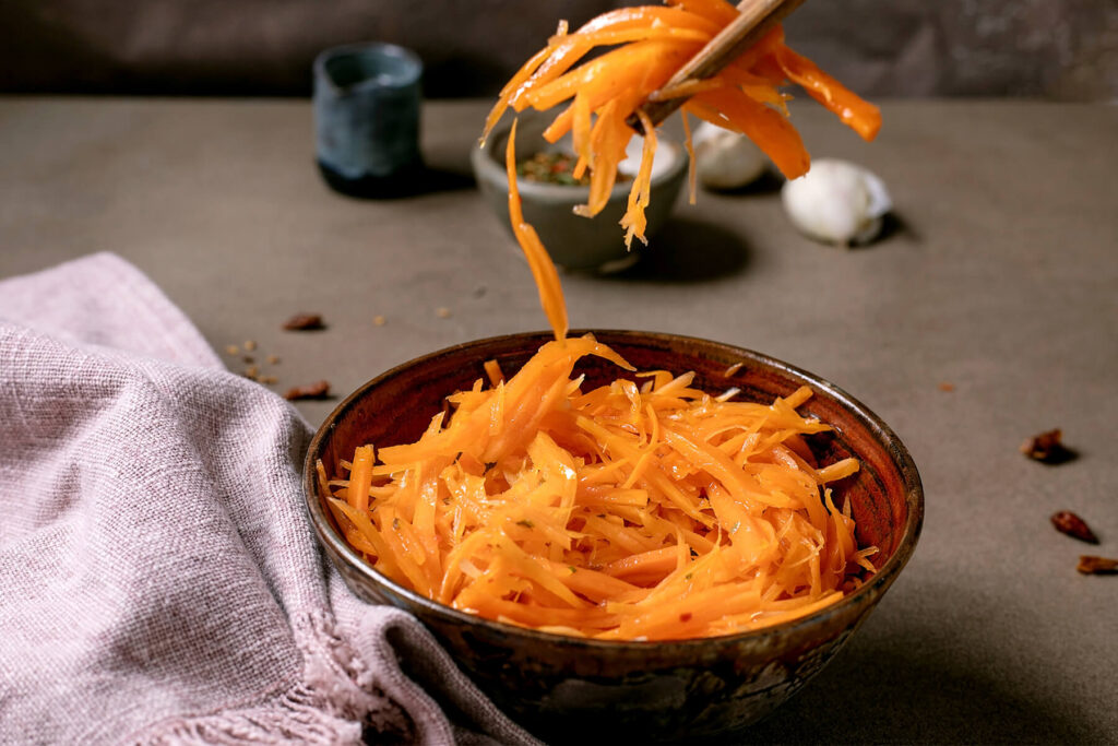 Koreanischer Karottensalat aus langen, glänzend marinierten Karottenstreifen in einer rustikalen Keramikschale, mit Stäbchen angehoben