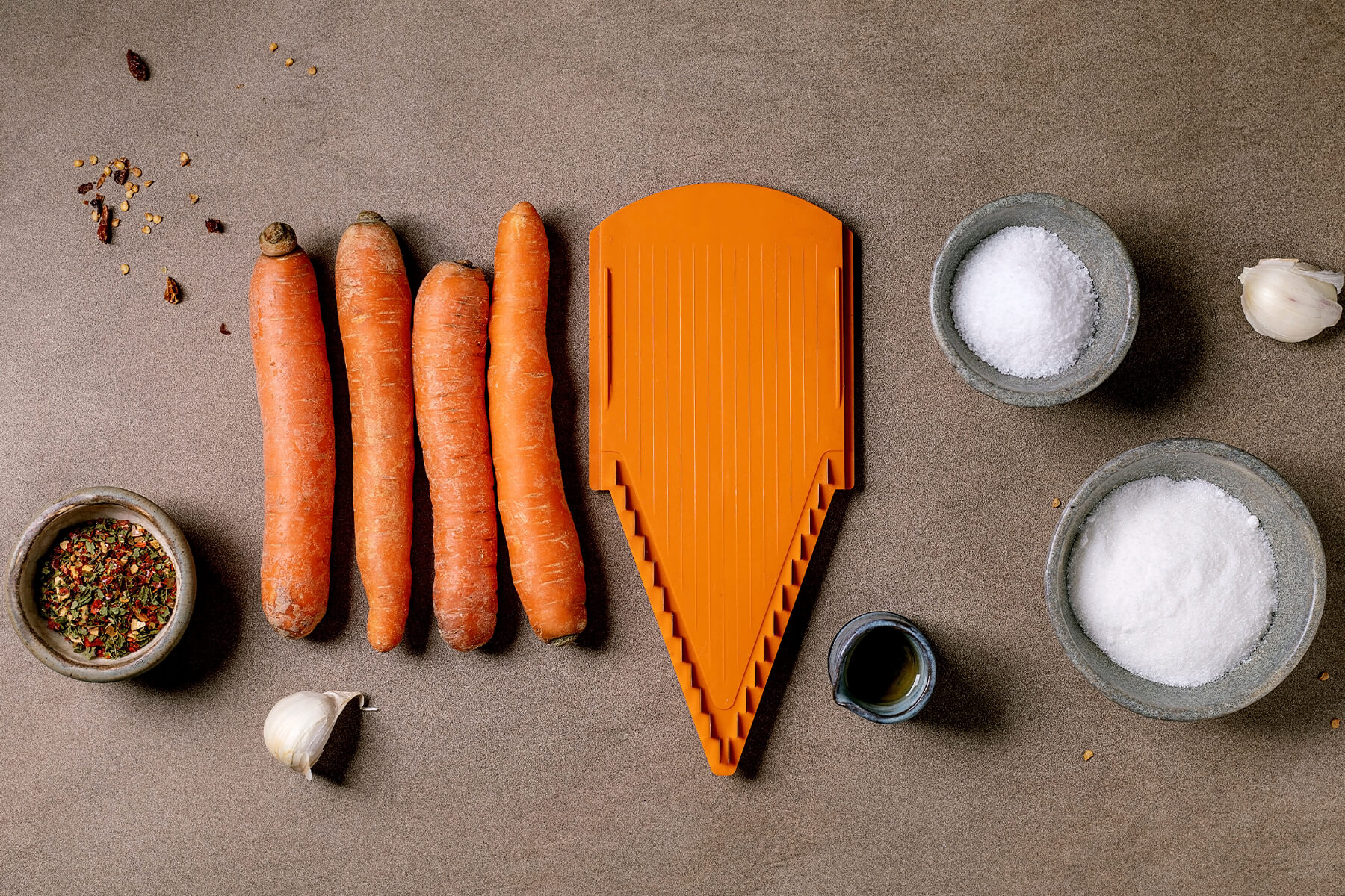 "Zutaten für koreanischen Karottensalat von oben: vier rohe Karotten neben einer orangefarbenen Julienne-Raspel, dazu Schälchen mit Salz, Knoblauchzehen und Gewürzen auf grauem Untergrund"