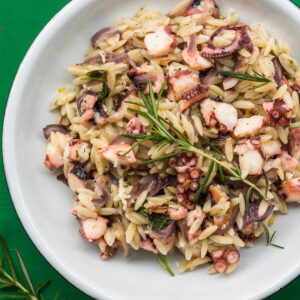 Kritharaki mit Oktopus, geschmorten roten Zwiebeln und frischem Rosmarin in einer weißen Schale auf grünem Untergrund