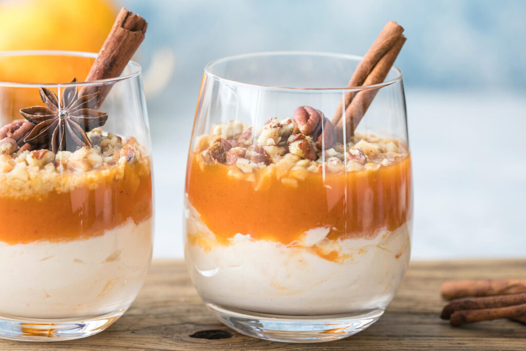 Zwei Gläser herbstliches Kürbis-Trifle mit heller Frischkäse-Creme, glänzender oranger Kürbisschicht und gehackten Pekannüssen, garniert mit Zimtstange und Sternanis