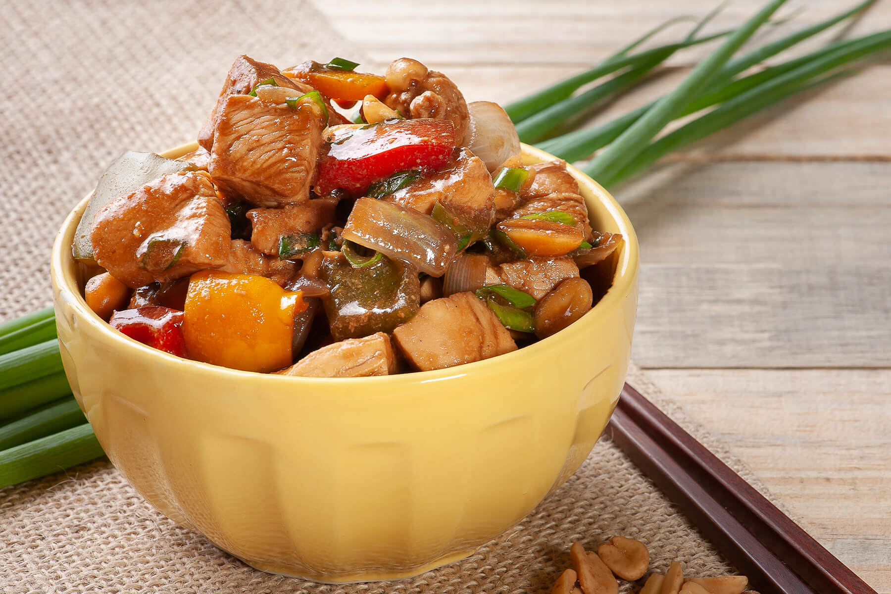 Schale mit Kung Pao Chicken von der Seite, glänzende Hähnchenwürfel mit roter und grüner Paprika, Zwiebeln und Frühlingszwiebeln, daneben Frühlingszwiebelstangen und Stäbchen