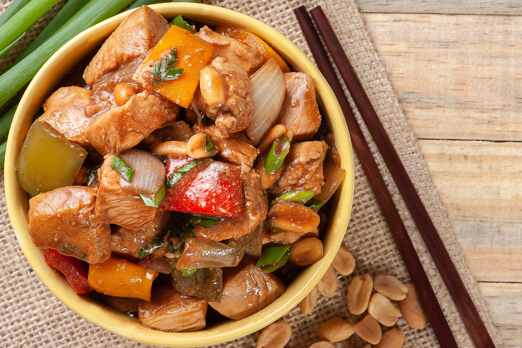 Kung Pao Chicken von oben in einer Schale, Hähnchenwürfel mit bunter Paprika, Zwiebeln, Erdnüssen und Frühlingszwiebeln, daneben Erdnüsse und Stäbchen