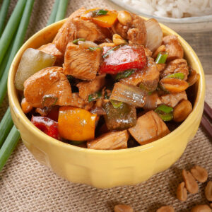 Kung Pao Chicken mit Hähnchenwürfeln, buntem Paprika, Zwiebeln, Frühlingszwiebeln und Erdnüssen in glänzender Sojasauce in einer gelben Schale, dazu Reis