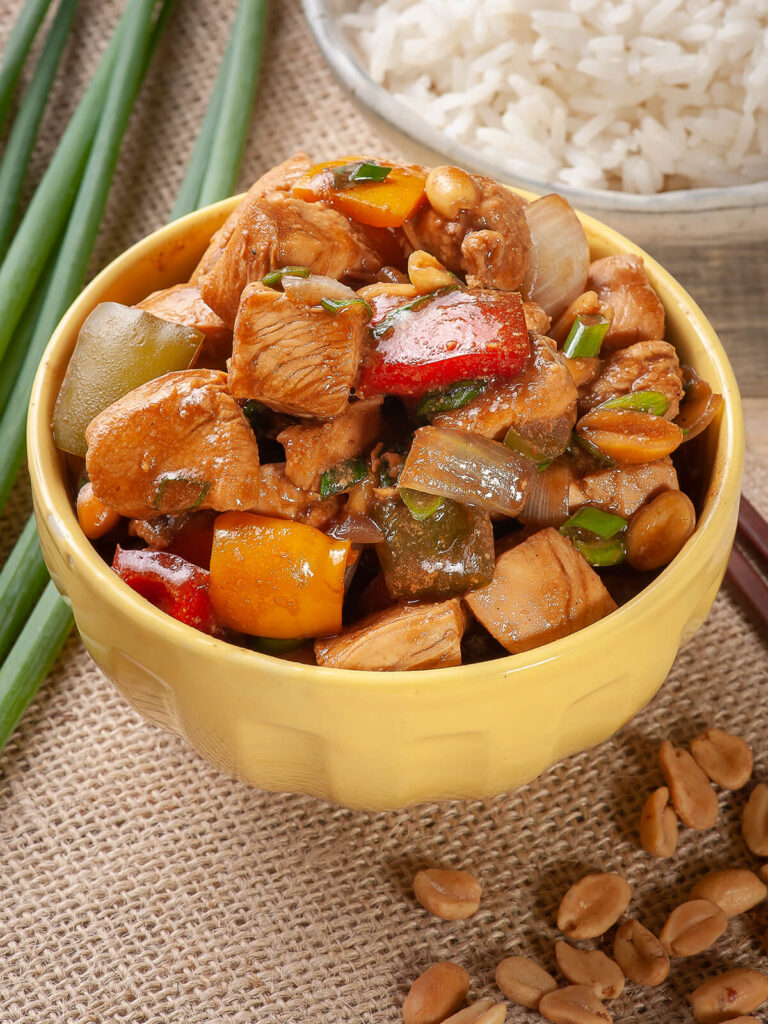 Kung Pao Chicken mit Hähnchenwürfeln, buntem Paprika, Zwiebeln, Frühlingszwiebeln und Erdnüssen in glänzender Sojasauce in einer gelben Schale, dazu Reis