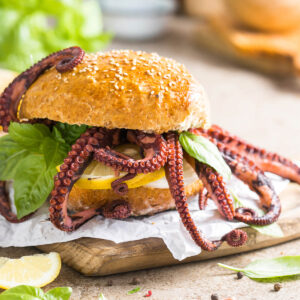 Oktopus-Burger im Sesambrötchen mit heraushängenden Tentakeln, Zitronenscheibe und Basilikum auf einem Holzbrett