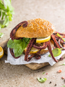 Fertig zusammengesetzter Oktopus-Burger im Vollkorn-Sesambrötchen mit heraushängenden Tentakeln, Zitronenscheiben und Basilikum auf Papier