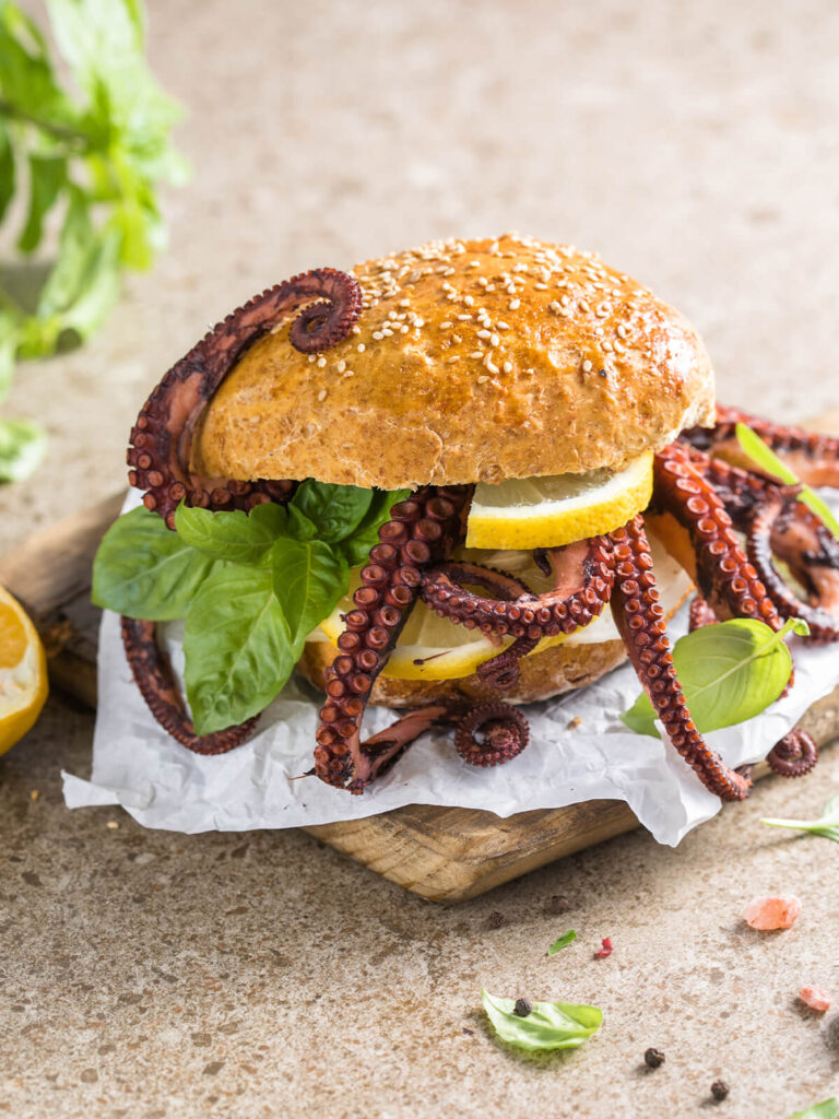 Fertig zusammengesetzter Oktopus-Burger im Vollkorn-Sesambrötchen mit heraushängenden Tentakeln, Zitronenscheiben und Basilikum auf Papier