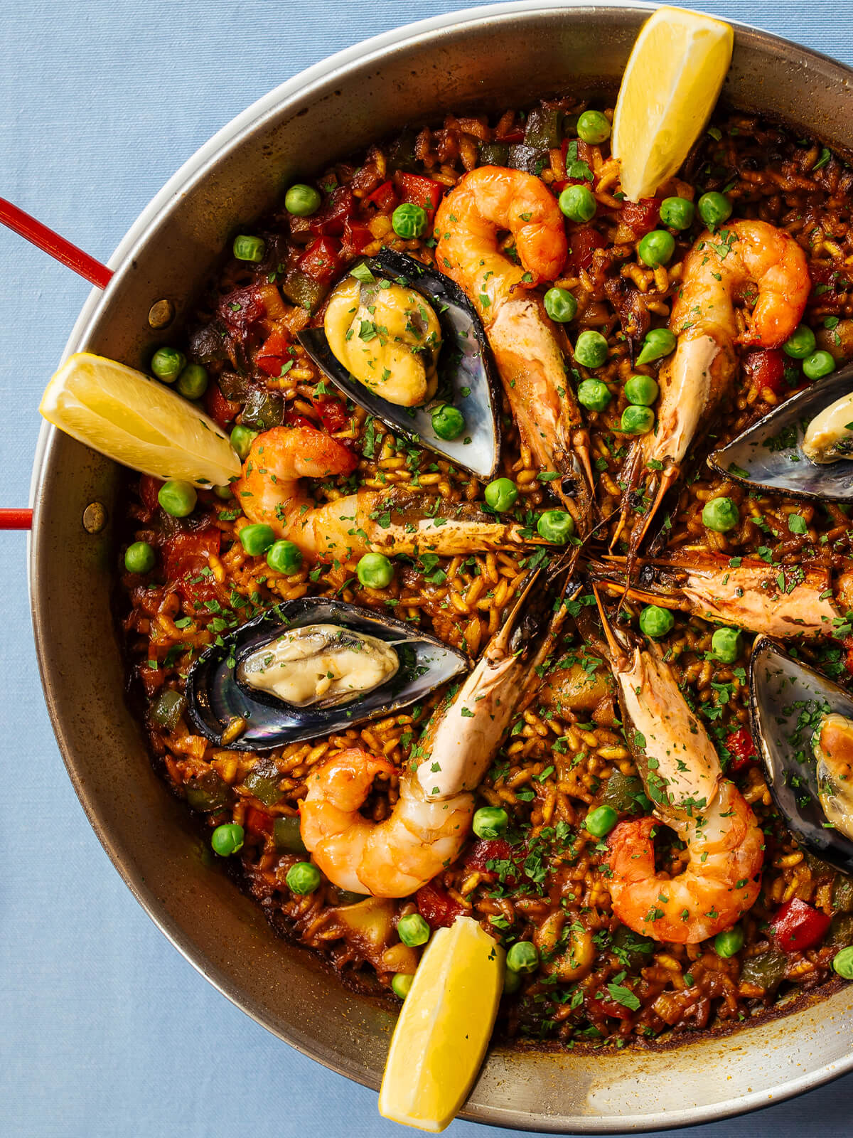 Spanische Paella de Marisco in der flachen Paella-Pfanne mit roten Griffen – safrangelber Reis mit ganzen Garnelen, Miesmuscheln in der Schale, grünen Erbsen und Zitronenspalten