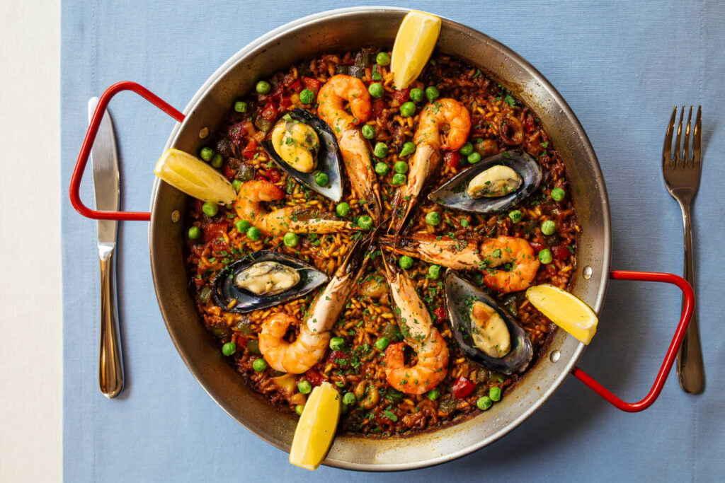 Spanische Paella de Marisco von oben in der flachen Paella-Pfanne mit roten Griffen – safrangelber Reis mit Garnelen, Miesmuscheln, grünen Erbsen und Zitronenspalten