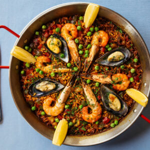 Spanische Paella de Marisco von oben in der flachen Paella-Pfanne mit roten Griffen – safrangelber Reis mit Garnelen, Miesmuscheln, grünen Erbsen und Zitronenspalten