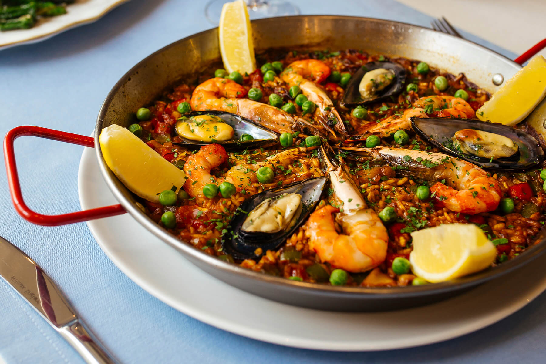 Seitliche Nahaufnahme der spanischen Meeresfrüchte-Paella mit Garnelen, geöffneten Miesmuscheln und Zitronenspalten in der Pfanne auf gedecktem Tisch