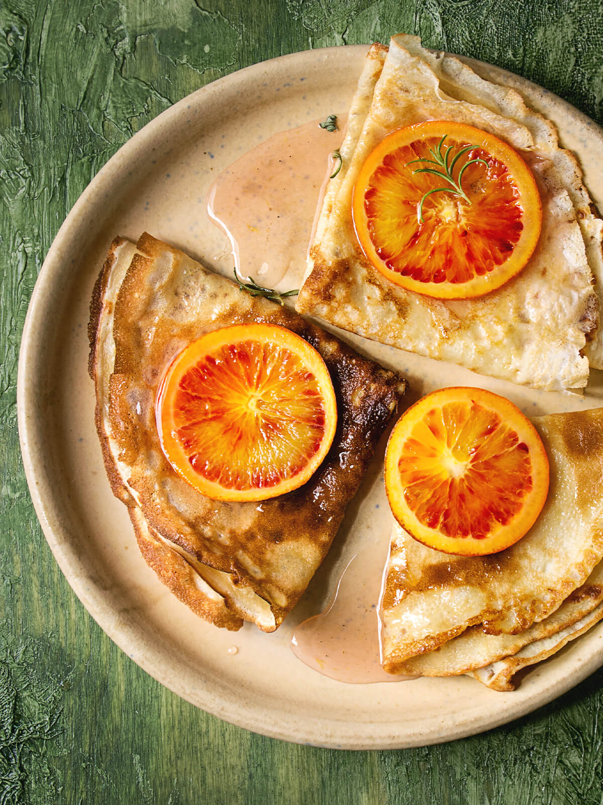 Crêpes Suzette mit Blutorangen auf einem hellen Teller, zu Dreiecken gefaltet, glänzend in einer karamellisierten Blutorangen-Butter-Sauce, mit Fruchtscheiben und einem Rosmarinzweig