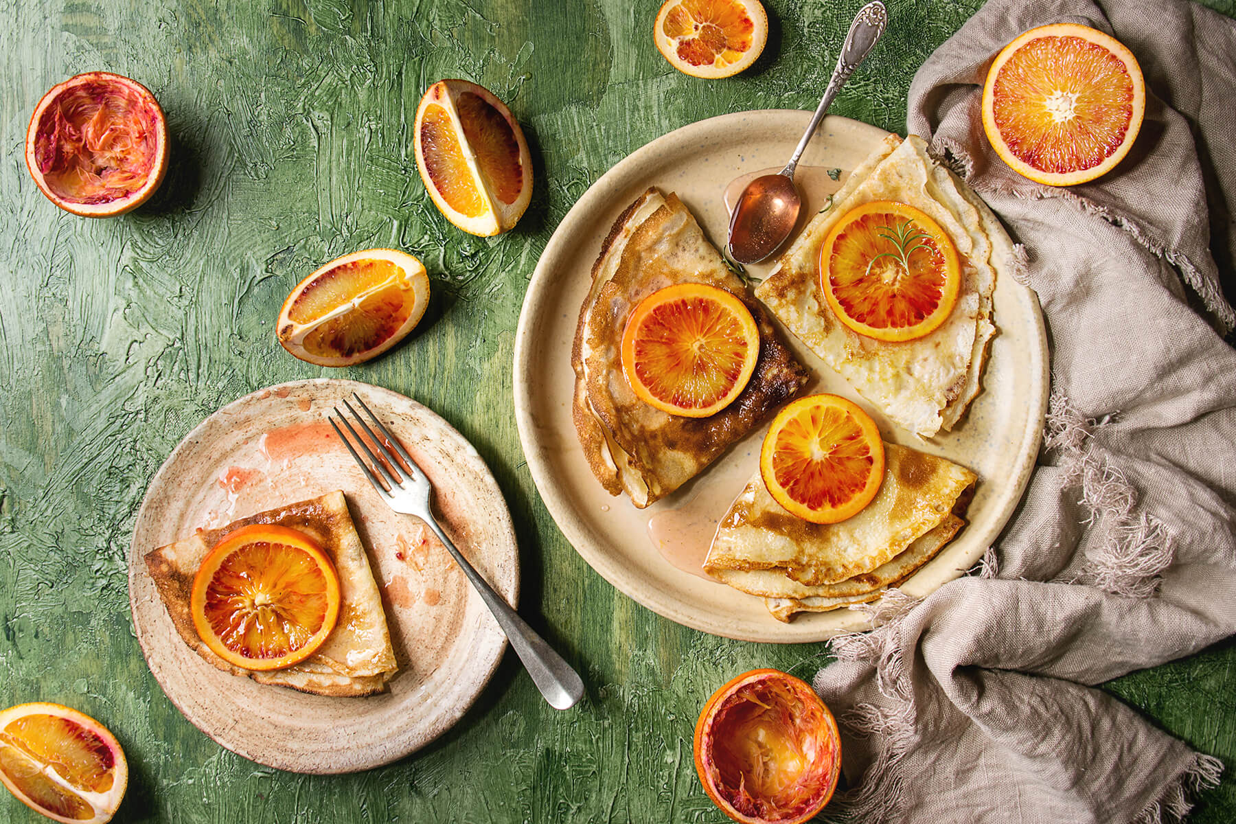 Zwei Teller mit dünnen Crêpes Suzette und Blutorangen, dazu die Blutorangen-Butter-Sauce, ein Löffel und halbierte Blutorangen auf grünem Hintergrund