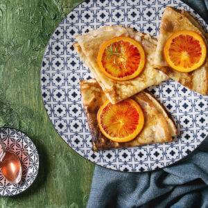 Crêpes Suzette mit Blutorangen, zu Dreiecken gefaltet auf einem gemusterten Teller, glänzend in der Blutorangen-Butter-Sauce, mit karamellisierten Scheiben und Rosmarin