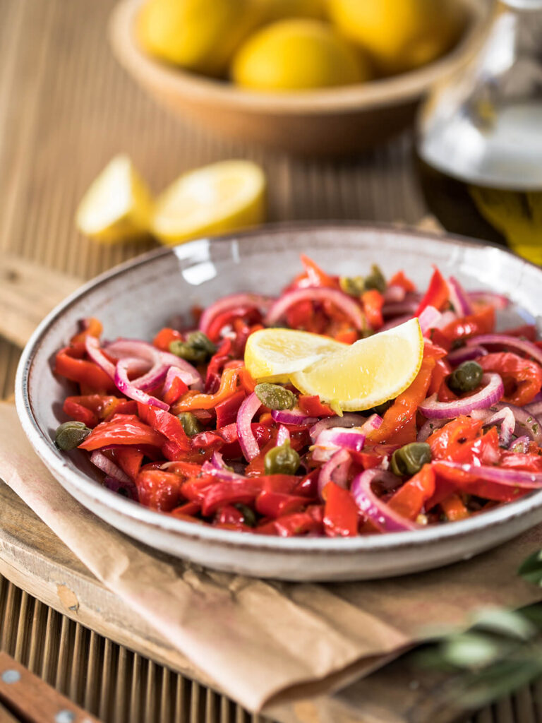 Warmer Paprikasalat aus gerösteten roten Paprikastreifen mit roten Zwiebelringen, großen Kapern und zwei Zitronenspalten in einer hellen Schale