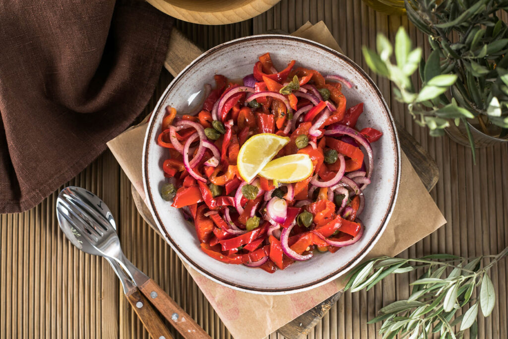 Warmer Paprikasalat mit roten Zwiebelringen, großen Kapern und zwei Zitronenspalten in einer hellen Schale, von oben fotografiert