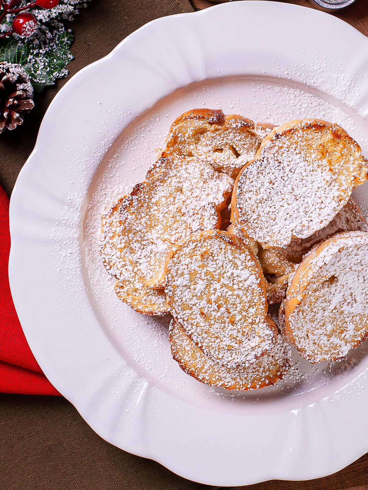 Rabanadas - goldbraun gebratene, runde Brotscheiben dick mit Puderzucker bestäubt auf einem weißen Teller mit weihnachtlicher Deko