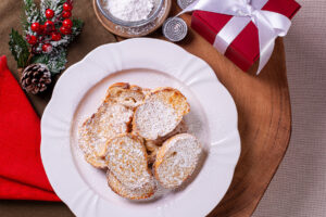 Rabanadas mit Puderzucker bestäubt auf einem weißen Teller, daneben ein Sieb mit Puderzucker, ein Teelicht und ein rotes Weihnachtsgeschenk