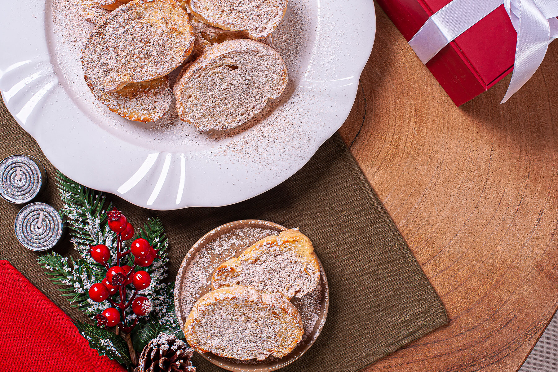 Rabanadas auf einem weißen Teller und auf einem kleinen Beistellteller, mit einer Mischung aus Puderzucker und Zimt bestäubt, daneben ein rotes Weihnachtsgeschenk