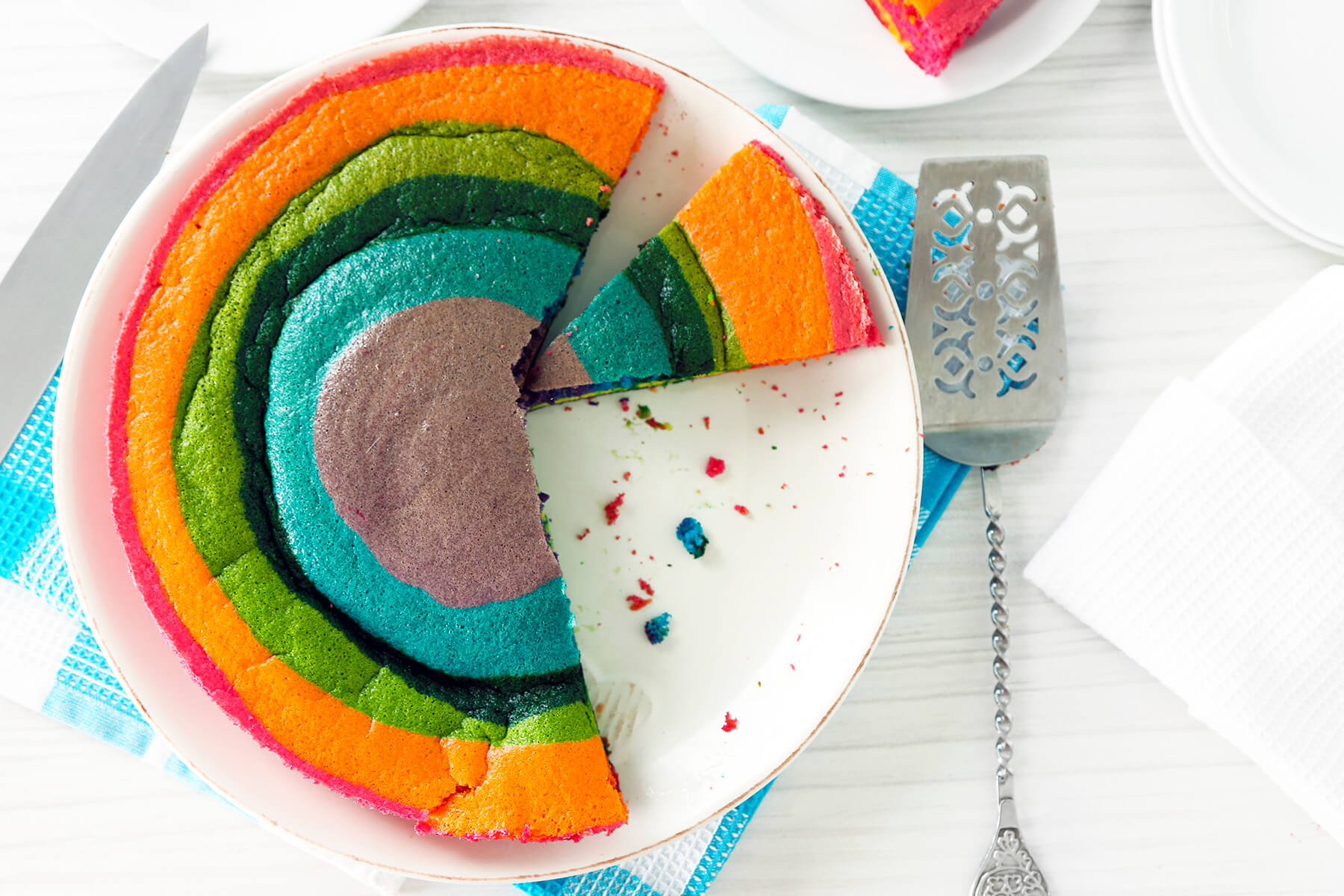 Regenbogen-Biskuit von oben mit herausgeschnittenem Stück, die konzentrischen Ringe und ein Tortenheber sind zu sehen