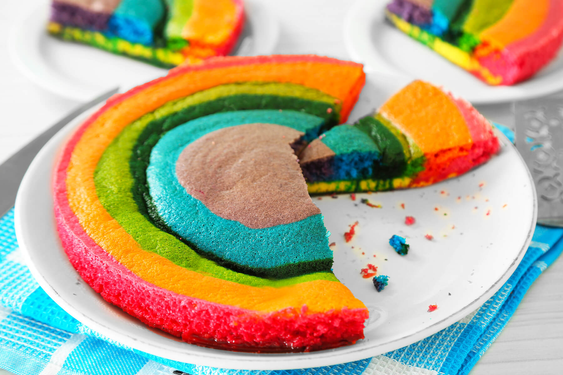 Angeschnittener Regenbogen-Biskuit auf einem Teller, ein Stück ist herausgenommen, daneben liegen Krümel und der Tortenheber
