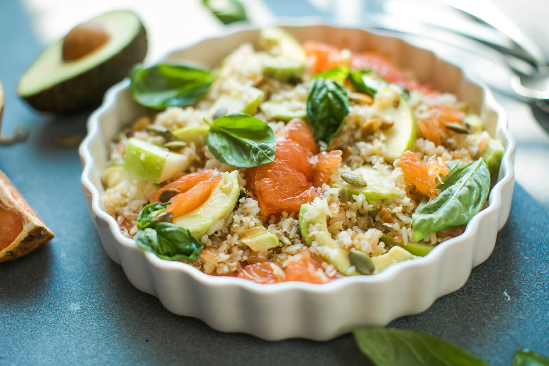Reissalat mit Grapefruit von der Seite in einer flachen weißen geriffelten Form, mit rosa Grapefruit-Filets, grünen Apfelspalten, Avocadoscheiben, gerösteten Kürbiskernen und Basilikumblättern