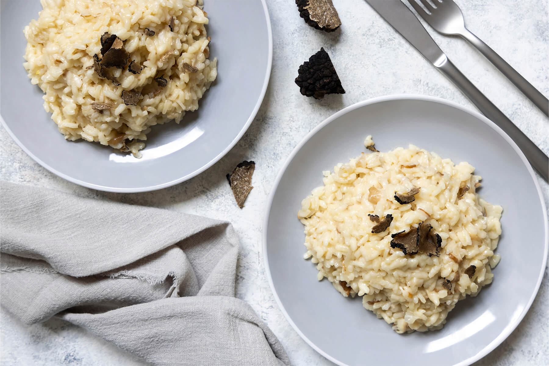 Zwei graue Teller mit cremigem Steinpilz-Risotto von oben fotografiert, mit gehobelten schwarzen Trüffelscheiben und Trüffelstücken daneben