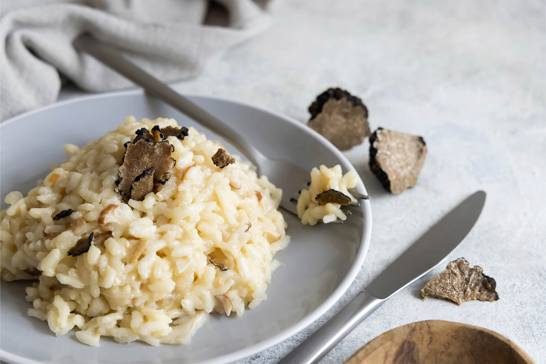 Cremiges Steinpilz-Risotto auf einem grauen Teller mit gehobelten schwarzen Trüffelscheiben, eine Gabel am Rand und Trüffelstücke daneben