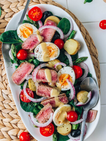 Salade Niçoise mit Thunfisch-Steak auf weißer Platte – rosa angebratenes Thunfisch-Steak, halbierte Eier, kleine Kartoffeln, Kirschtomaten, schwarze Oliven und rote Zwiebel auf jungem Spinat mit cremiger Dijon-Vinaigrette