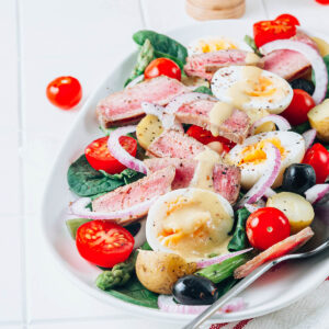 Salade Niçoise mit Thunfisch-Steak von oben – rosa angebratener Thunfisch, halbierte Eier, kleine Kartoffeln, Kirschtomaten, schwarze Oliven und rote Zwiebel auf jungem Spinat mit Dijon-Vinaigrette