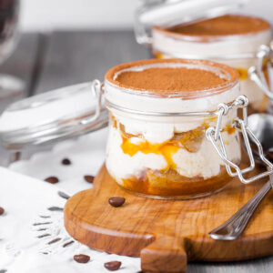 Zwei offene Bügelgläser mit Sanddorn-Tiramisu auf einem Holzbrett, sichtbare Schichten aus Mascarpone-Creme, getränktem Biskuit und orangem Sanddorn-Püree