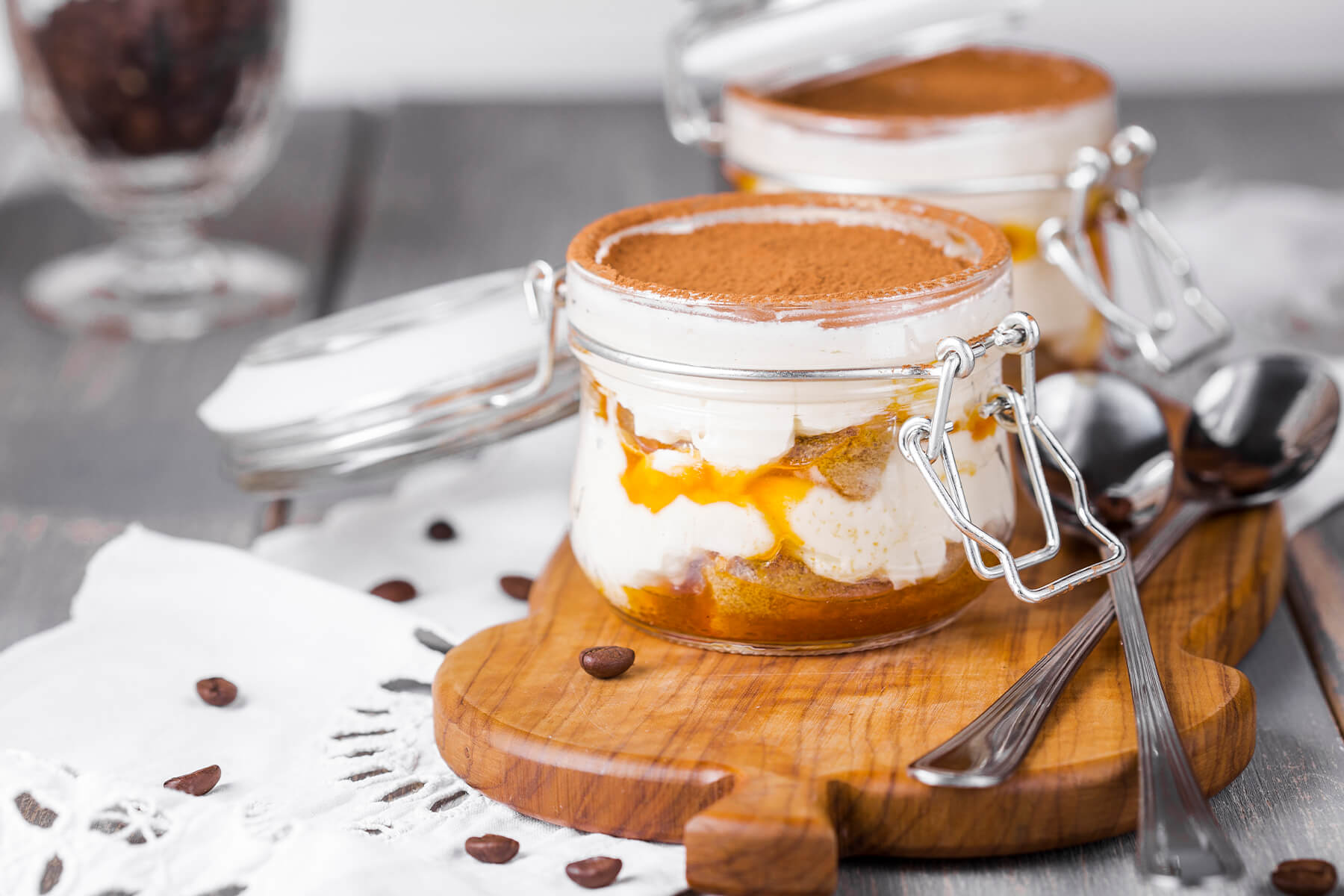 Zwei offene Bügelgläser mit Sanddorn-Tiramisu auf einem Holzbrett, sichtbare Schichten aus Mascarpone-Creme, in Saft getränktem Biskuit und orangem Sanddorn-Püree, Kaffeebohnen nur als Deko daneben