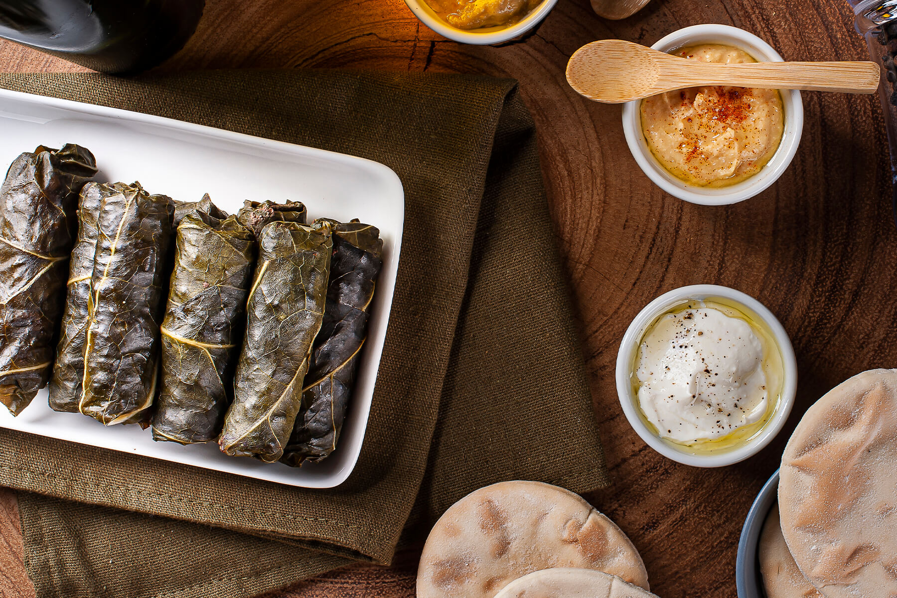 Gefüllte Weinblätter von oben auf einem weißen Teller, daneben Hummus, ein Joghurt-Dip mit Pfeffer und Olivenöl sowie Fladenbrot