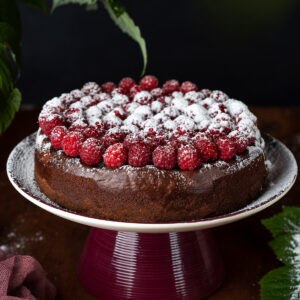 Runde Schokoladen-Trüffeltorte ohne Mehl mit Mandeln auf einem beerenroten Tortenständer, der hohe Rand ohne Glasur, die Oberfläche mit frischen Himbeeren in konzentrischen Ringen belegt und mit Puderzucker bestäubt
