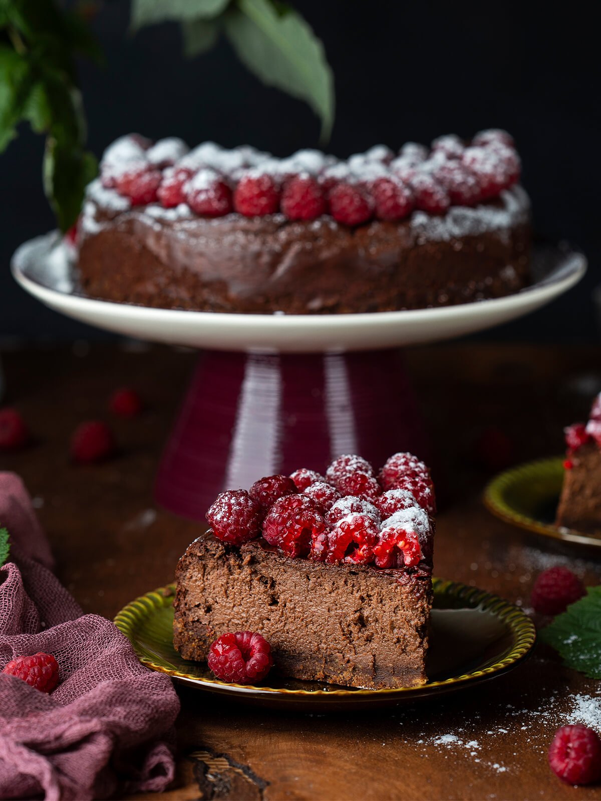 Angeschnittene Schokoladen-Trüffeltorte ohne Mehl, ein Stück auf einem grünen Teller zeigt die dichte, porenfreie und milchschokoladenbraune Krume ohne separaten Boden, darüber eine dünne dunkle Schokoglasur und Himbeeren