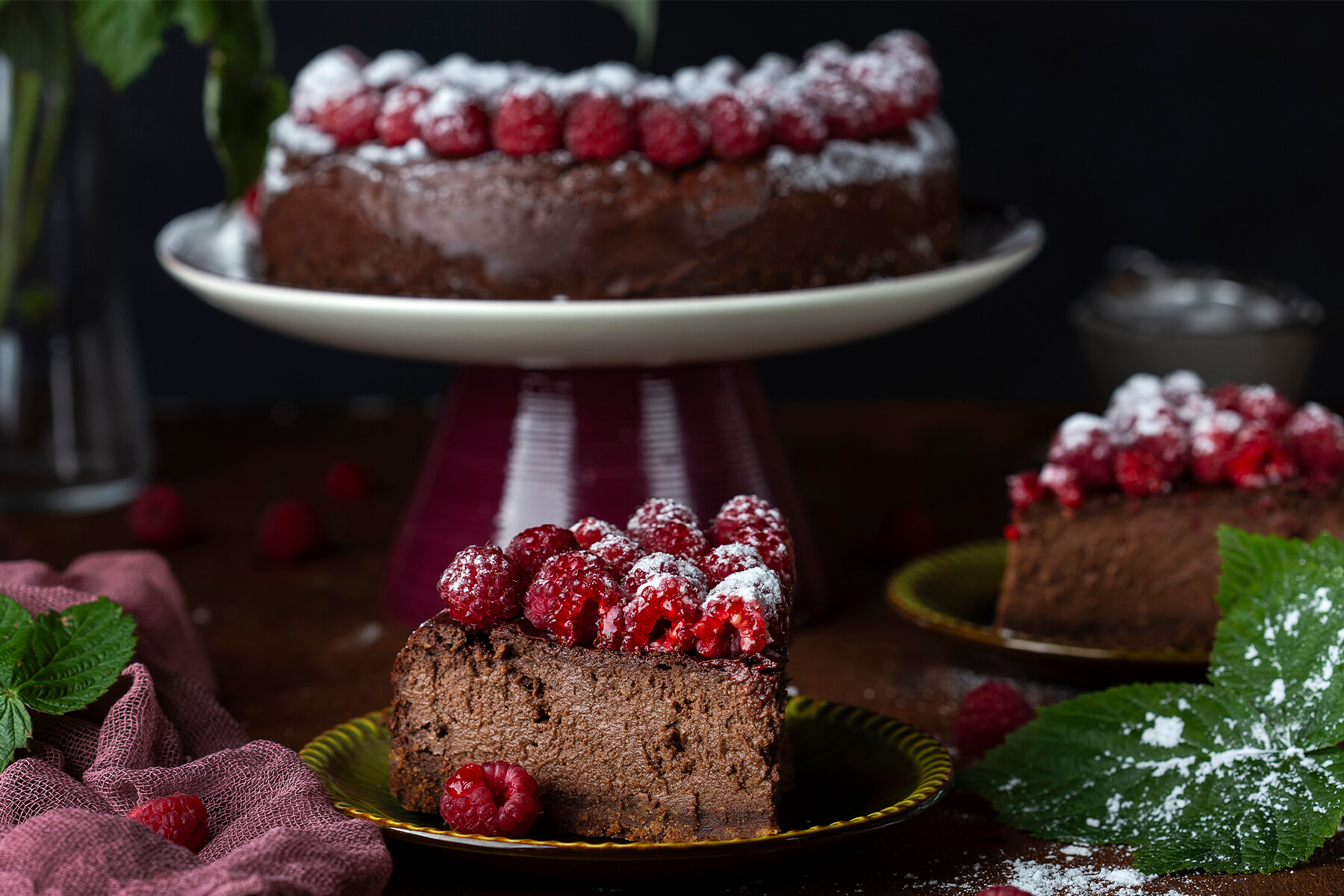 Zwei Stücke der Schokoladen-Trüffeltorte ohne Mehl mit Mandeln auf grünen Tellern, die Schnittflächen zeigen eine gleichmäßig dichte Krume ohne Poren, dahinter die angeschnittene Torte auf dem Tortenständer