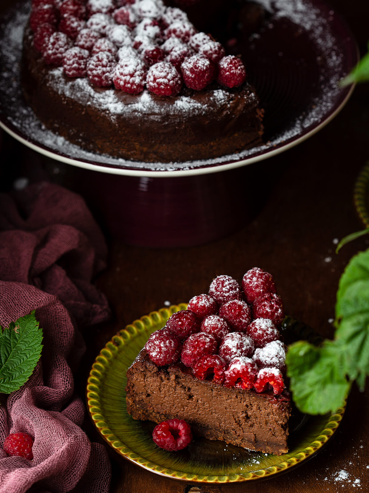 Ein Stück der Schokoladen-Trüffeltorte ohne Mehl mit Mandeln auf einem grünen Teller, die samtige Krume und die dünne Glasur gut sichtbar, im Hintergrund die angeschnittene Torte auf dem Tortenständer