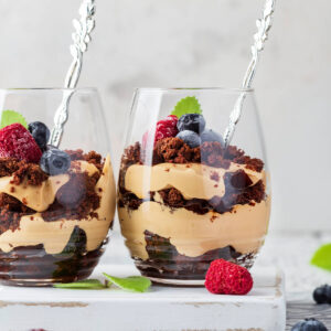 Zwei Gläser mit Schoko-Karamell-Schichtdessert, sichtbare Schichten aus dunklen Schokobröseln und karamellfarbener Creme, oben Himbeeren, Heidelbeeren und Minze