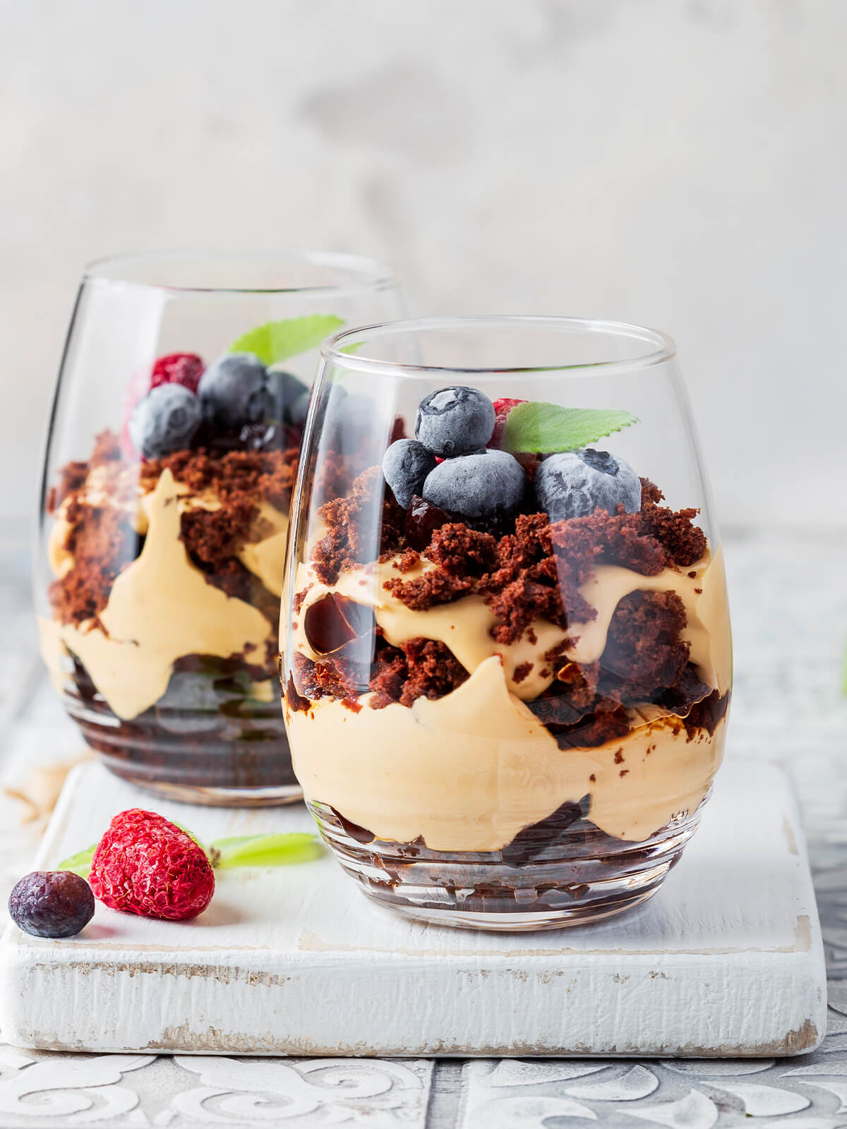 Zwei Gläser Schoko-Karamell-Schichtdessert mit Löffeln, oben üppig mit Himbeeren und Heidelbeeren belegt, dazwischen ein Minzblatt