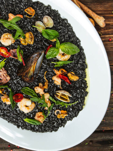Schwarzes Risotto mit Meeresfrüchten auf weißem Teller – tiefschwarzer cremiger Reis mit Miesmuscheln, Vongole, Garnelen, Tintenfisch, Rucola und Basilikum