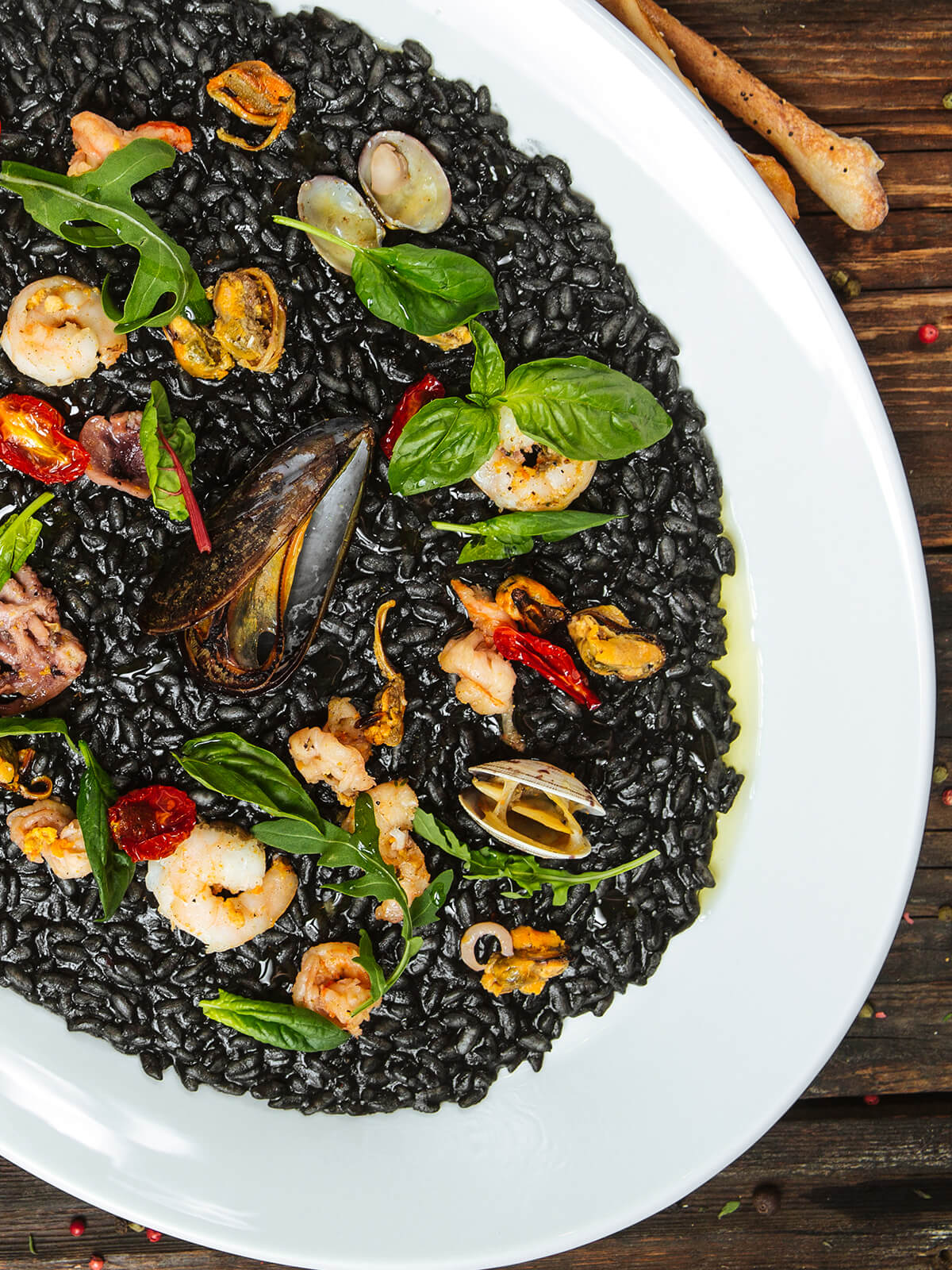 Schwarzes Risotto mit Meeresfrüchten auf weißem Teller – tiefschwarzer cremiger Reis mit Miesmuscheln, Vongole, Garnelen, Tintenfisch, Rucola und Basilikum