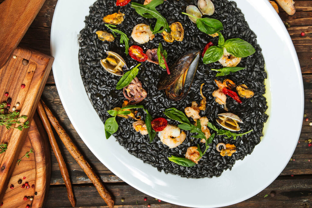 Schwarzes Risotto mit Meeresfrüchten auf weißem Teller von oben, garniert mit Miesmuscheln, Vongole, Garnelen, Tintenfisch, Rucola und Basilikum