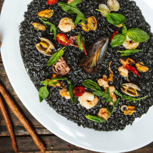 Schwarzes Risotto mit Meeresfrüchten auf weißem Teller von oben, garniert mit Miesmuscheln, Vongole, Garnelen, Tintenfisch, Rucola und Basilikum