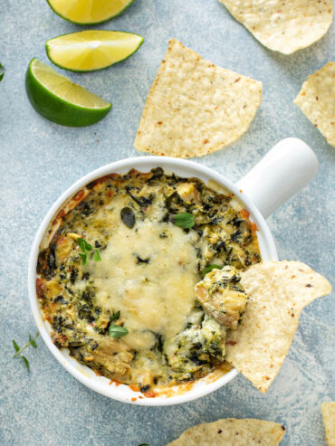 Warmer Spinat-Artischocken-Dip in weißer Auflaufform mit goldener Käsekruste, dazu Limettenspalten und Tortilla-Chips, von oben fotografiert