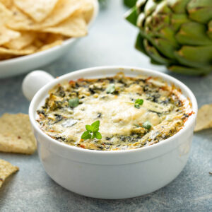 Warmer Spinat-Artischocken-Dip in weißer Auflaufform mit goldener Käsekruste, dahinter eine ganze Artischocke und ein Teller Tortilla-Chips