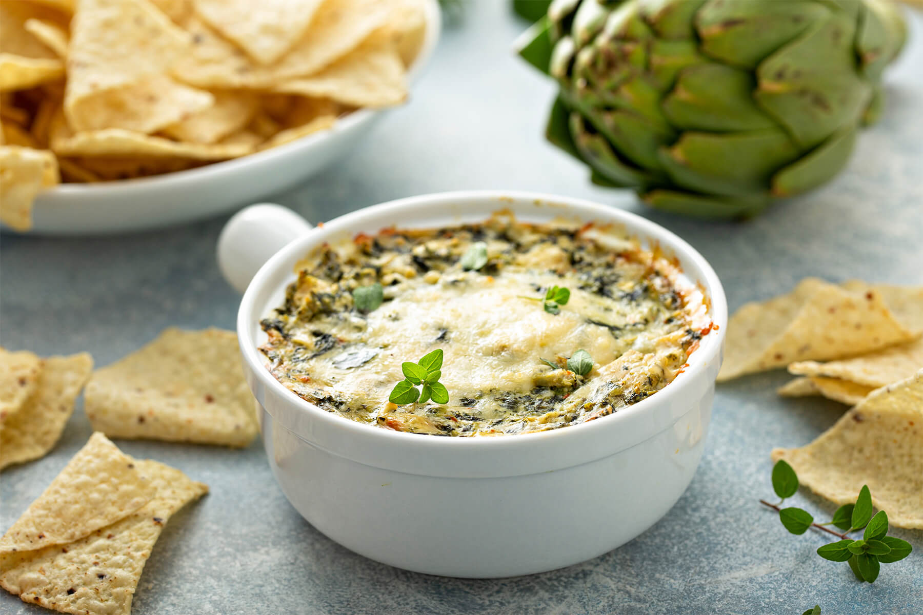 Warmer Spinat-Artischocken-Dip in weißer Auflaufform mit goldener Käsekruste, dahinter eine ganze Artischocke und ein Teller Tortilla-Chips