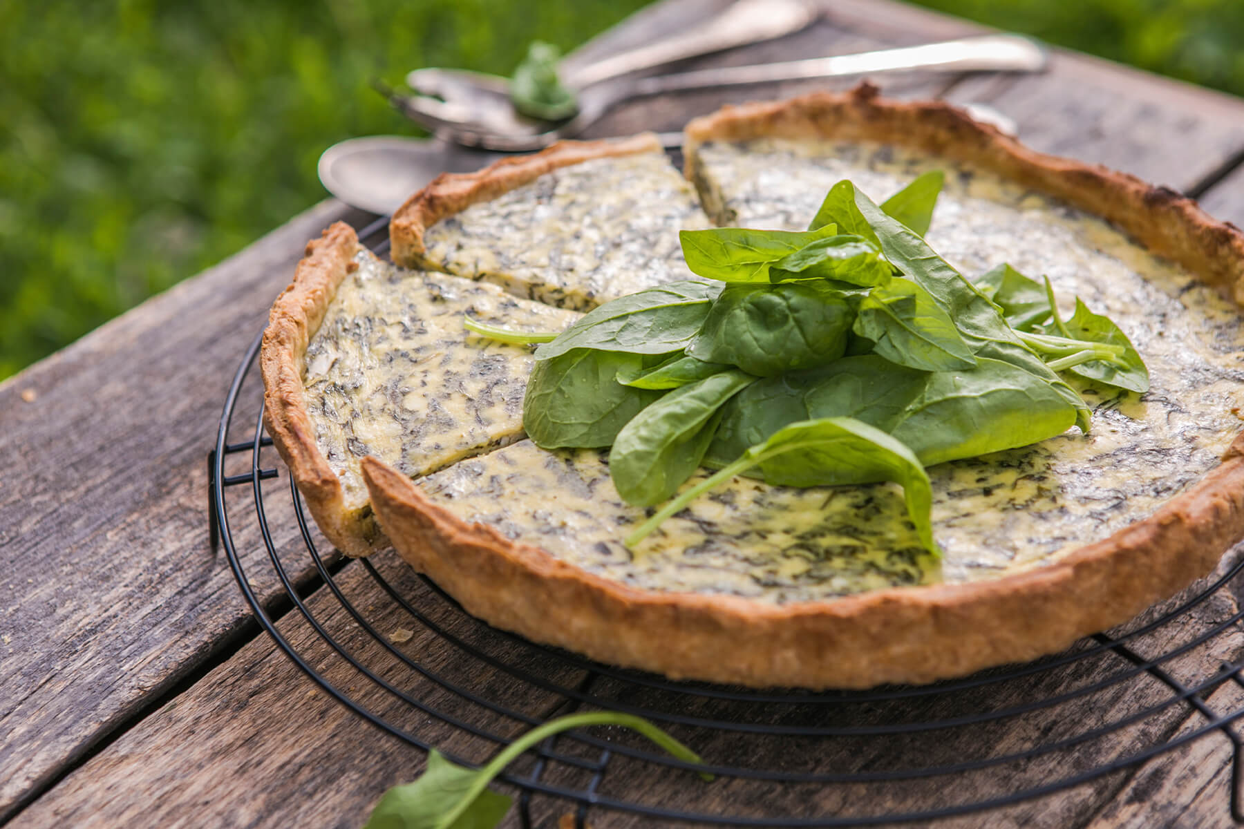 Runde Spinat-Quiche auf schwarzem Kuchengitter, in der Mitte mit einem Strauß frischer Spinatblätter belegt, auf verwittertem Holztisch im Garten