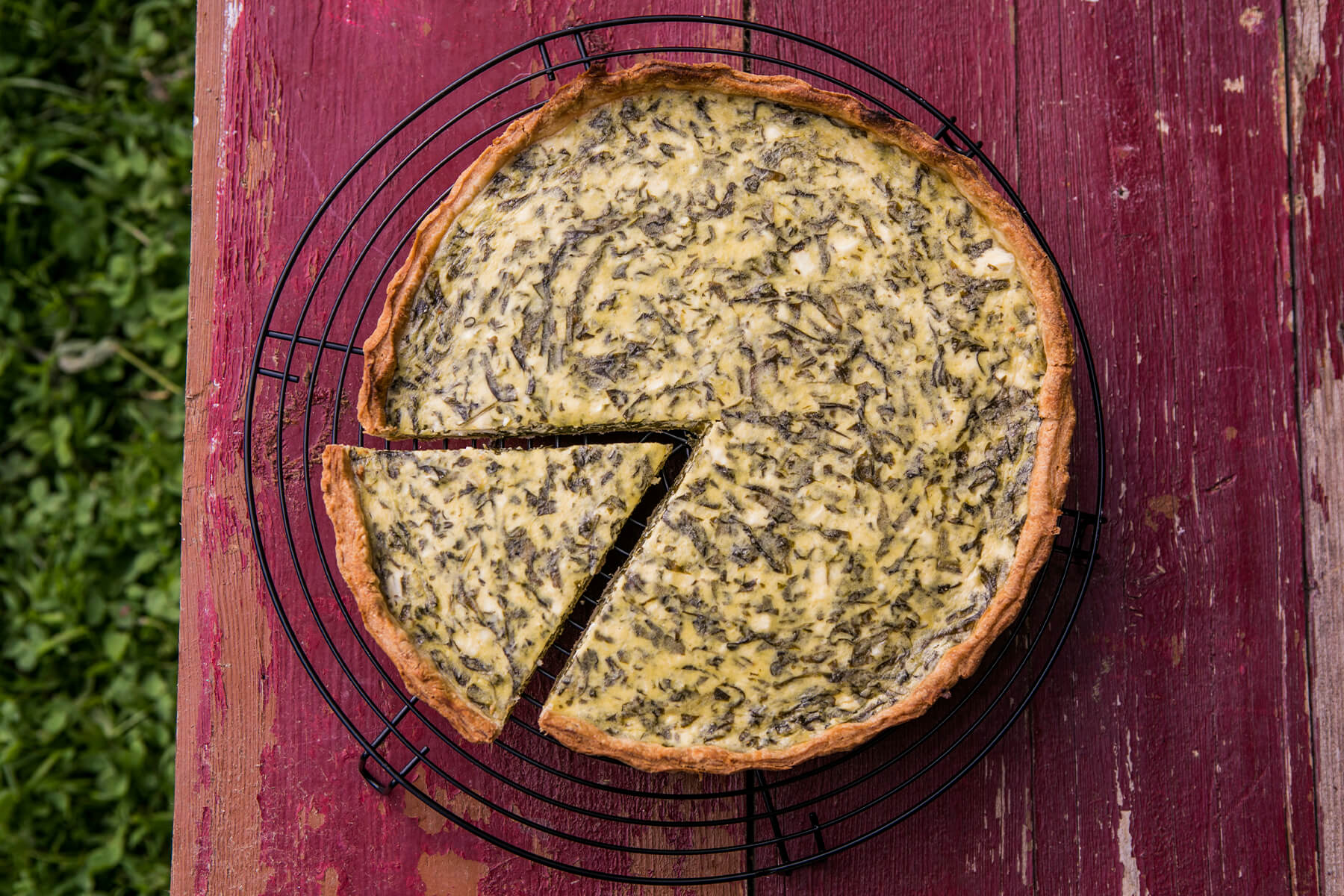Spinat-Quiche von oben auf schwarzem Kuchengitter, ein Stück bereits herausgeschnitten, mit goldgelb gebräuntem Rand und grün gesprenkelter Oberfläche